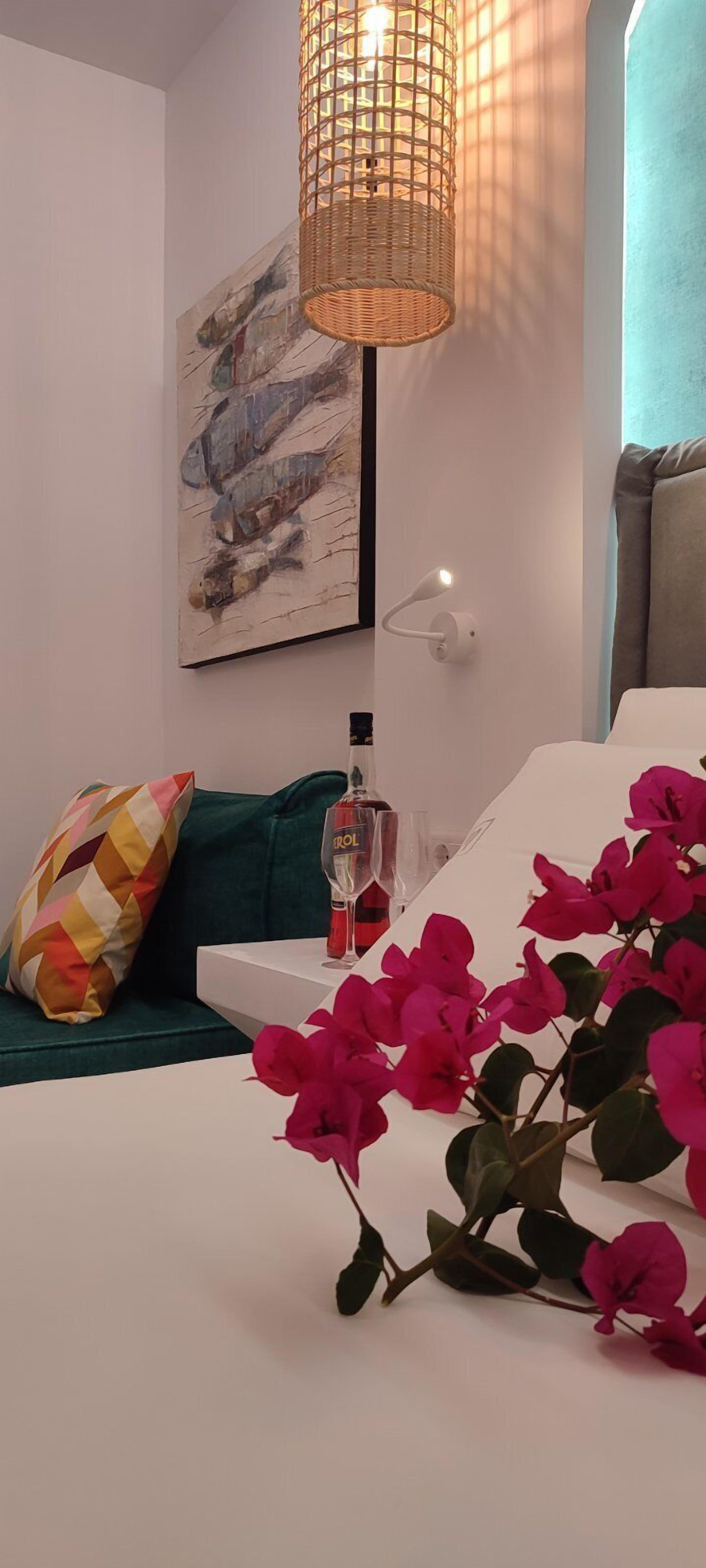 A modern living room with a green sofa, a colorful pillow, a white table with a bottle and glasses, and a vase of pink flowers.