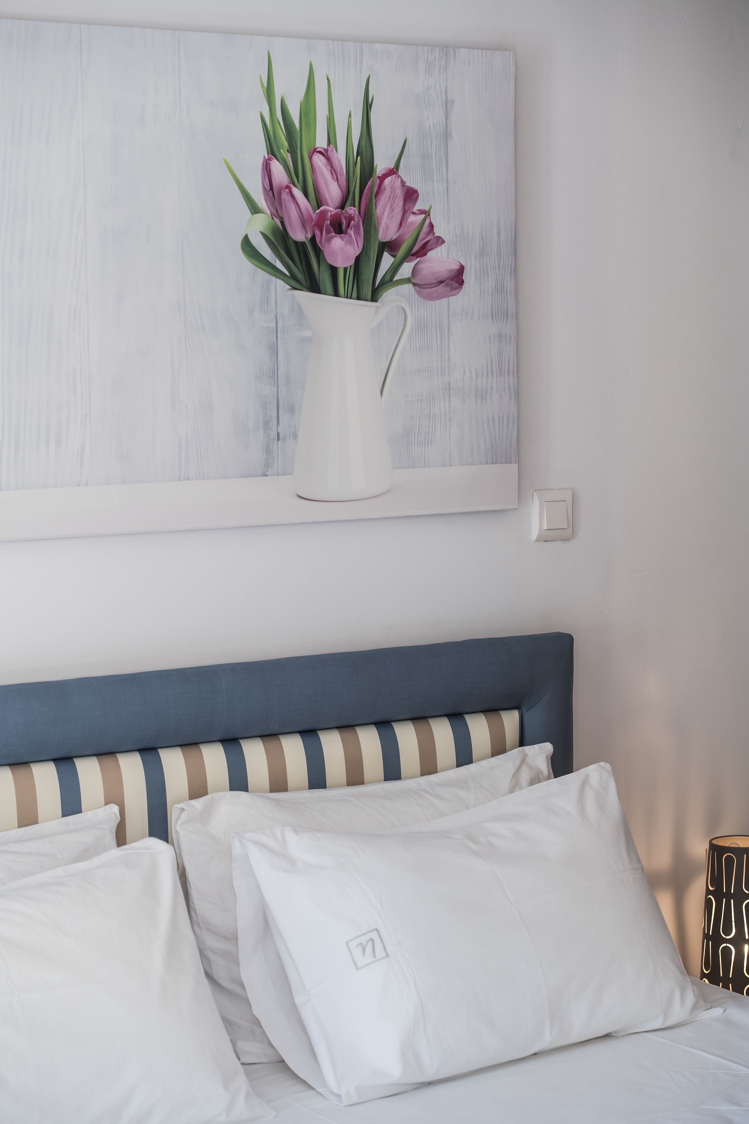 A bedroom with a bed featuring white and striped bedding, a blue headboard, and a painting of pink tulips on the wall.