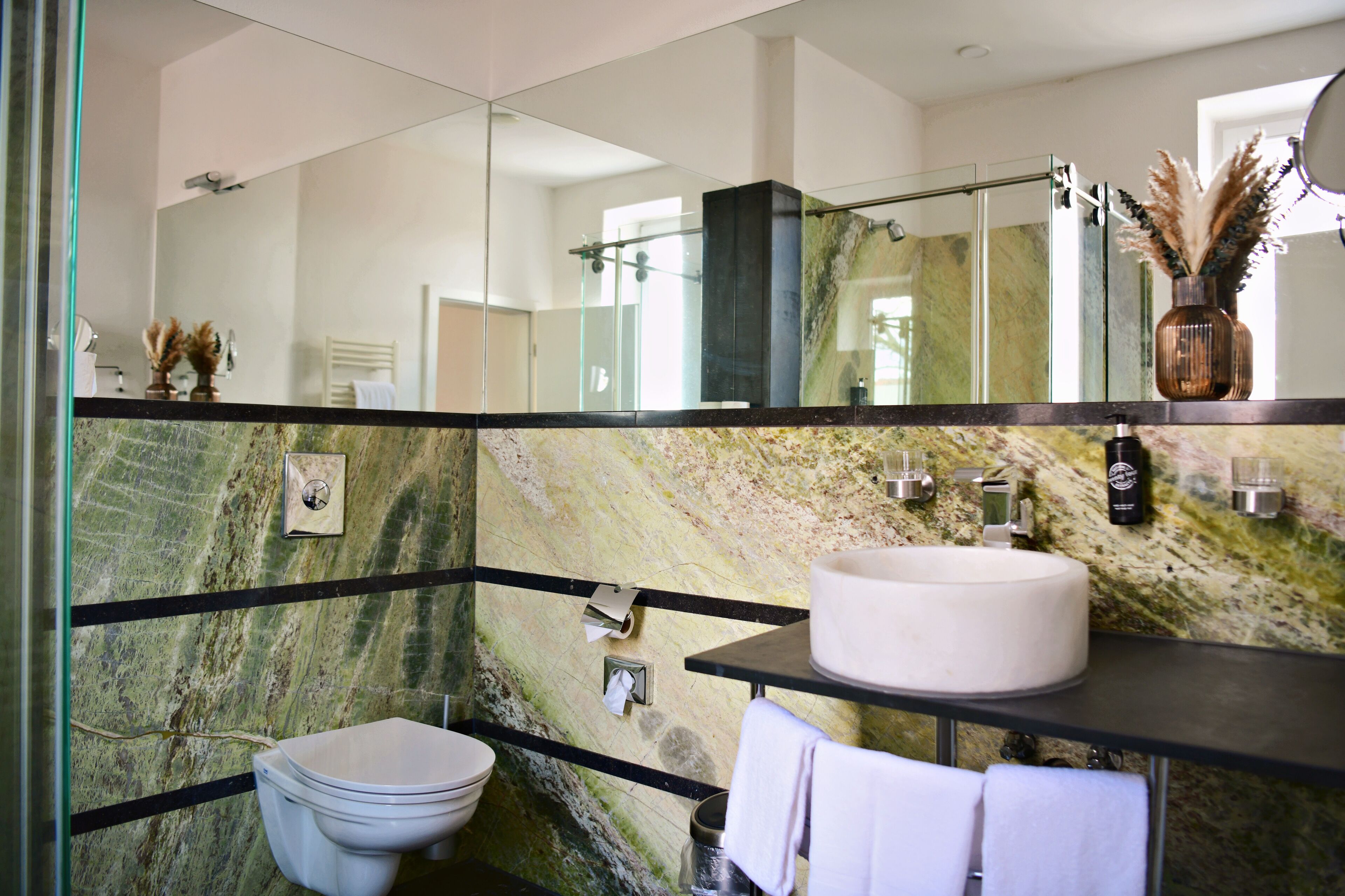 A modern bathroom with a marble wall, a round sink, a toilet, and a glass shower enclosure.