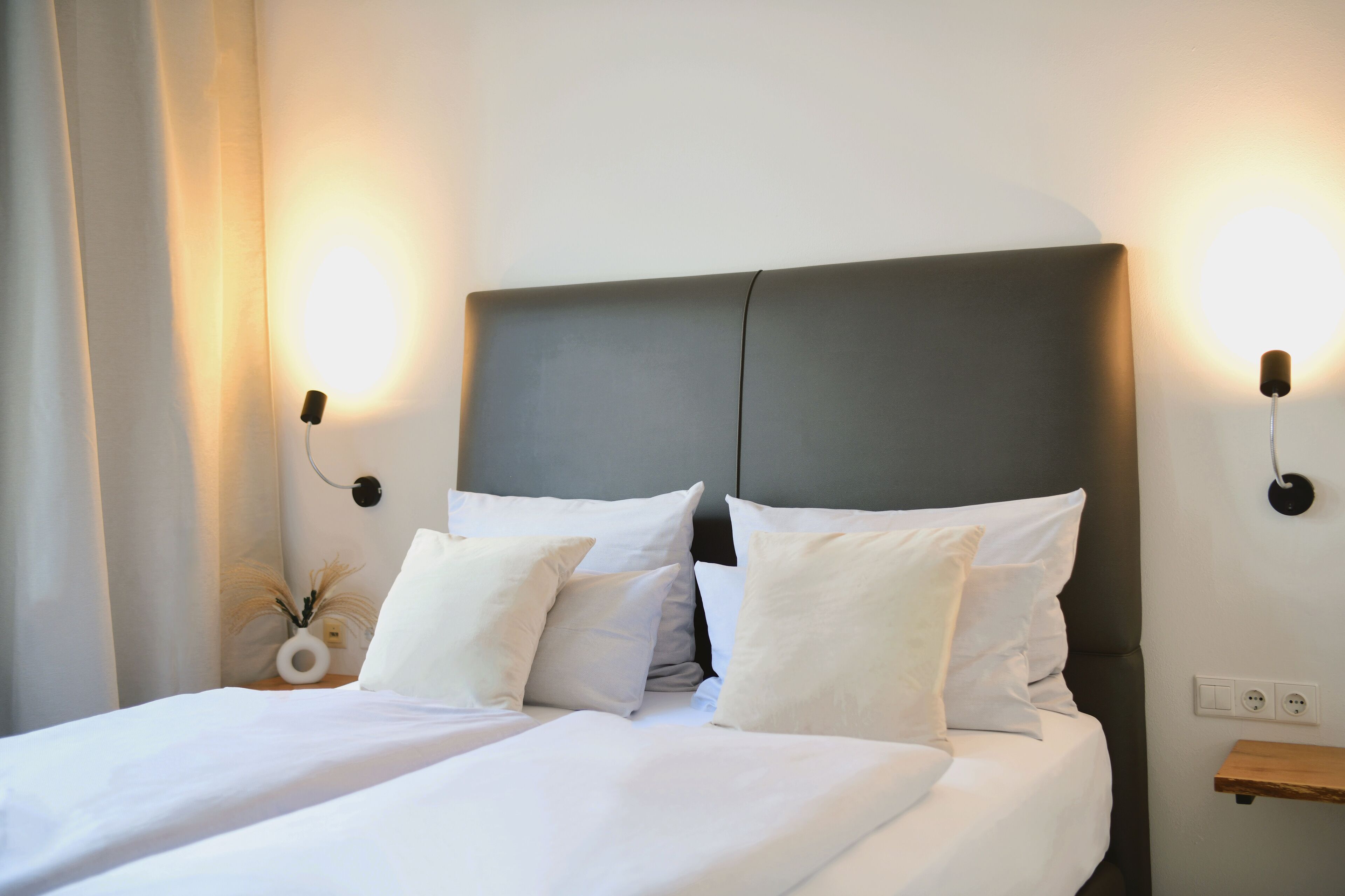 Una cama bien tendida con sábanas y almohadas blancas, un cabecero oscuro y dos lámparas fijadas a la pared a cada lado.