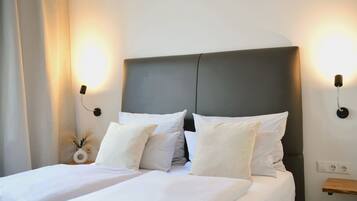 Una cama bien tendida con sábanas y almohadas blancas, un cabecero oscuro y dos lámparas fijadas a la pared a cada lado.