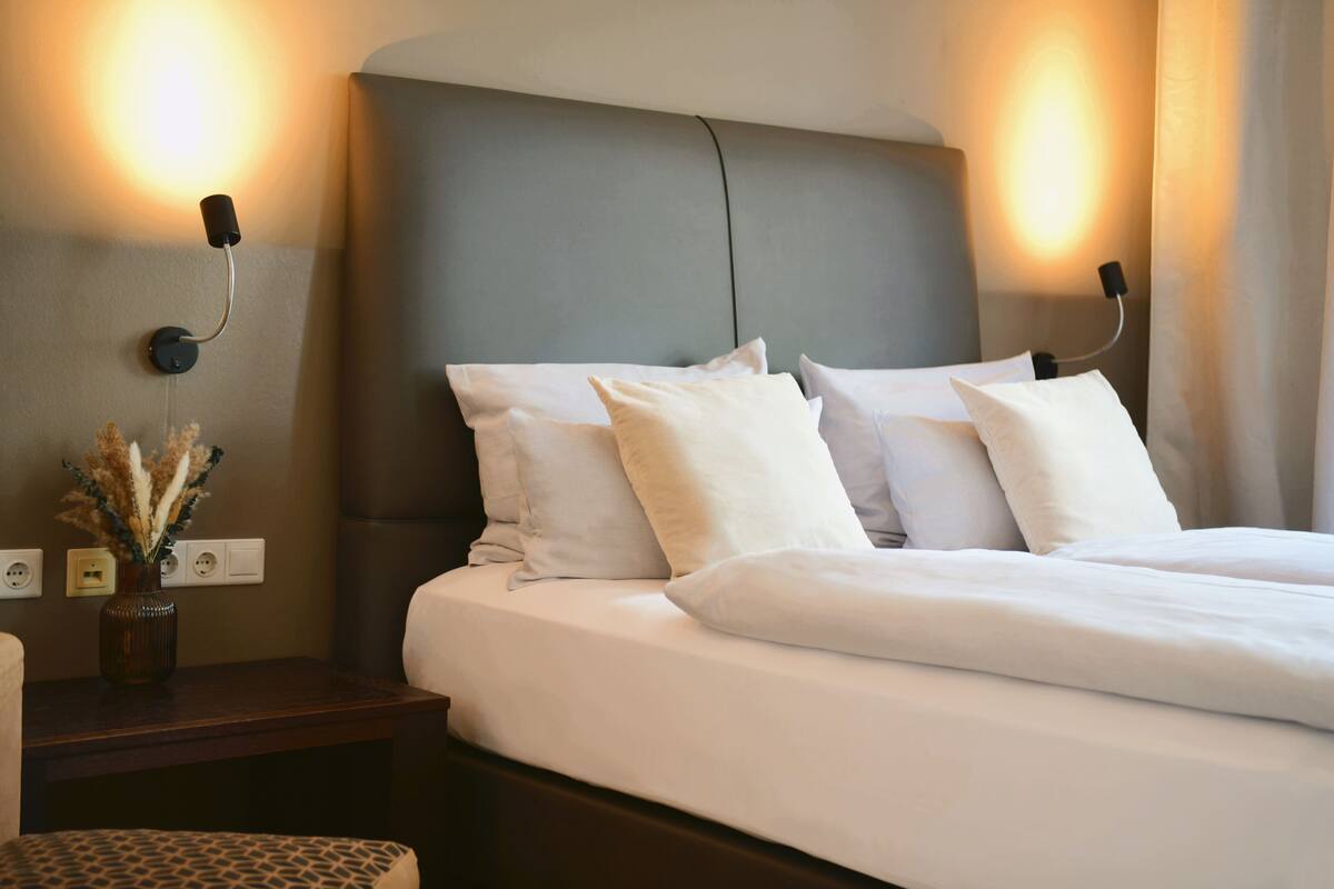 Una cama bien hecha con sábanas blancas y un cabecero gris, una mesita con un jarrón de flores secas y luces de lectura fijadas a la pared.
