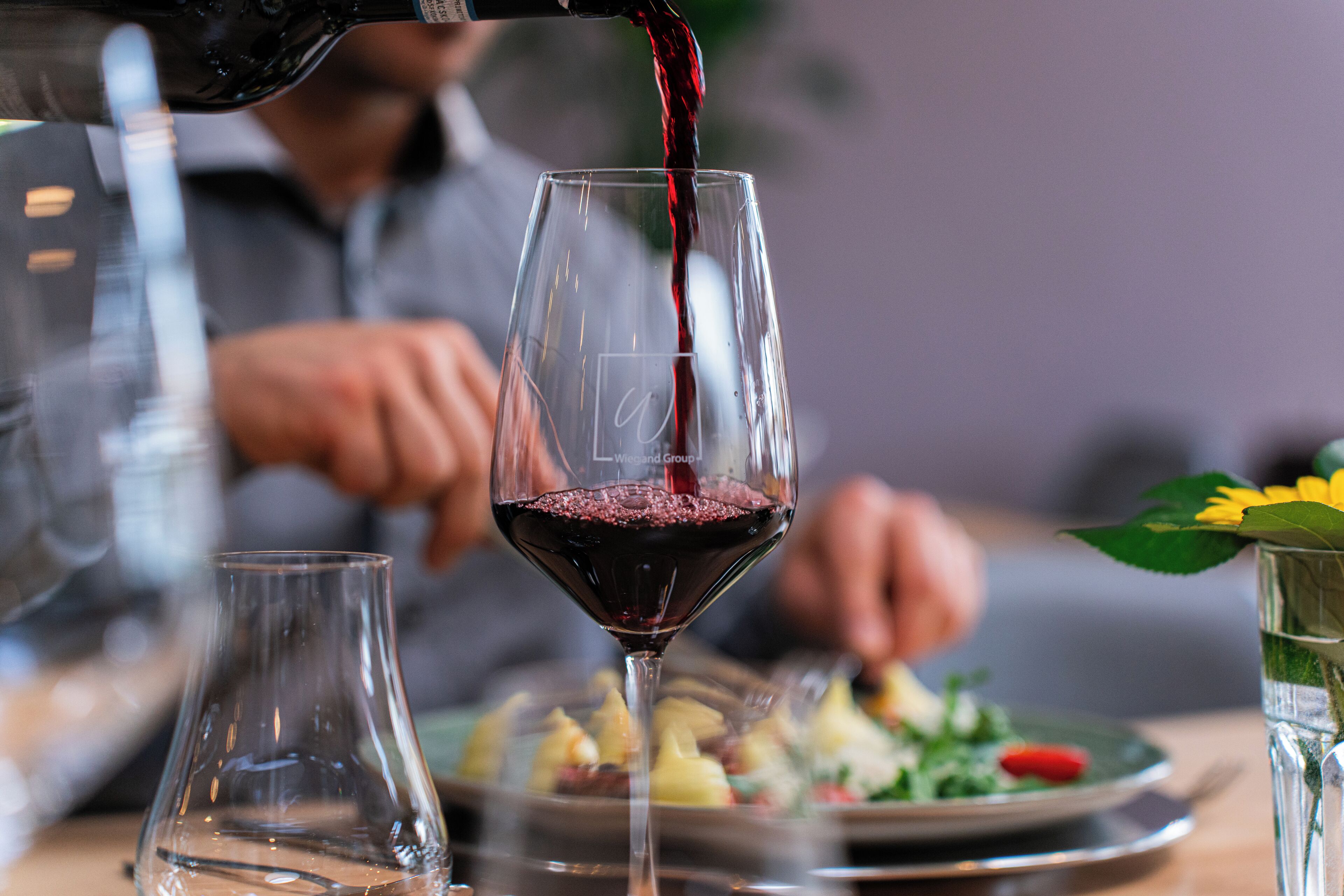A wine glass being filled with red wine, a clear glass, and a water glass with a flower on the table.