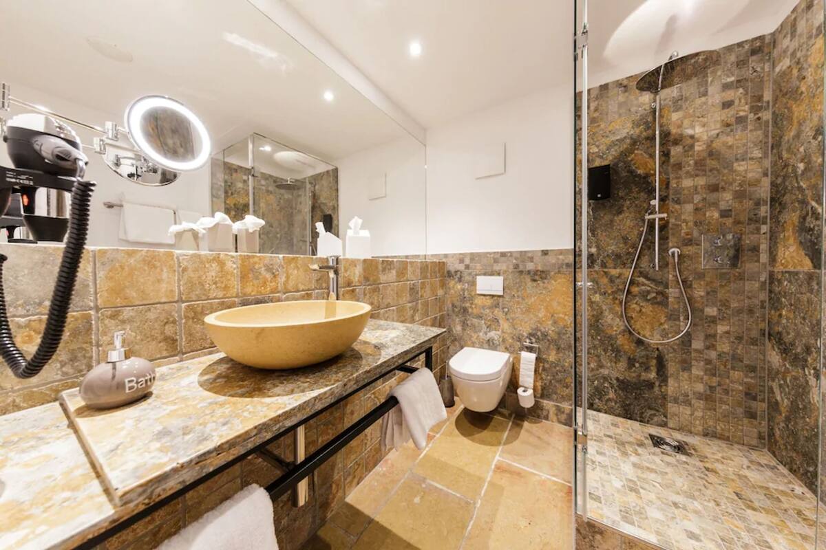 Un baño moderno con un lavamanos ovalado grande, un secador de pelo de mano, una ducha con puerta de vidrio y un muro con azulejos de mármol.