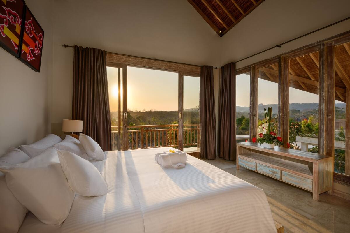 A spacious bedroom with a large bed, a wooden ceiling, and a balcony with a view of the landscape.