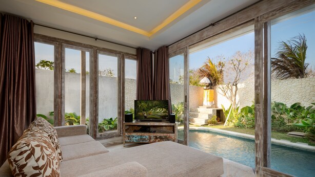 A modern living room with a large sofa, a television on a wooden stand, and a view of a swimming pool outside. - Adinda Balangan Beach Villa (Jimbaran)