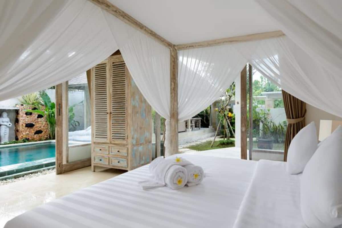 A bedroom with a large bed, white linens, and a canopy. There is a view of a pool and outdoor area with greenery.