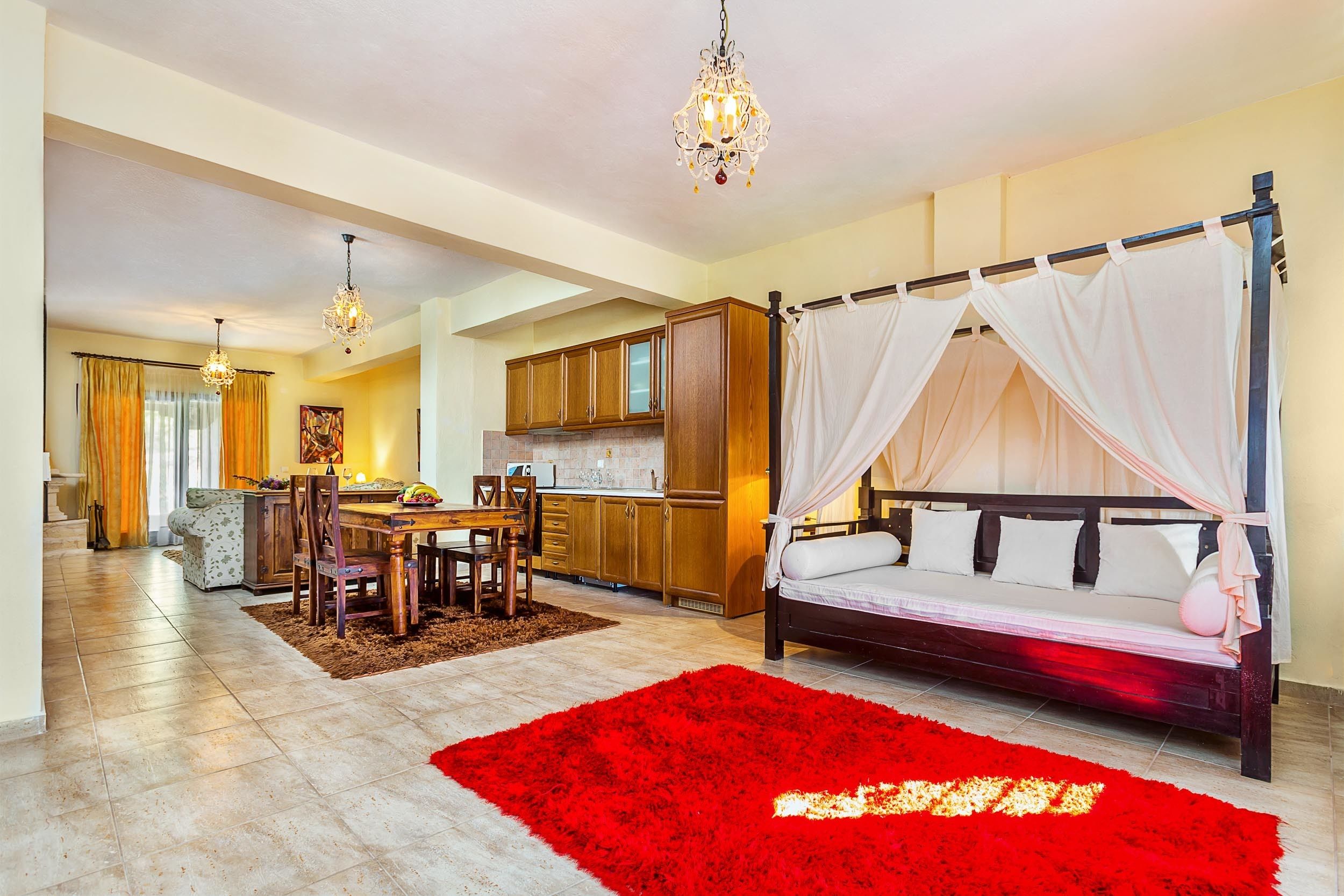 A spacious living area with a dining table, a large red rug, a four-poster bed, and a kitchen in the background.