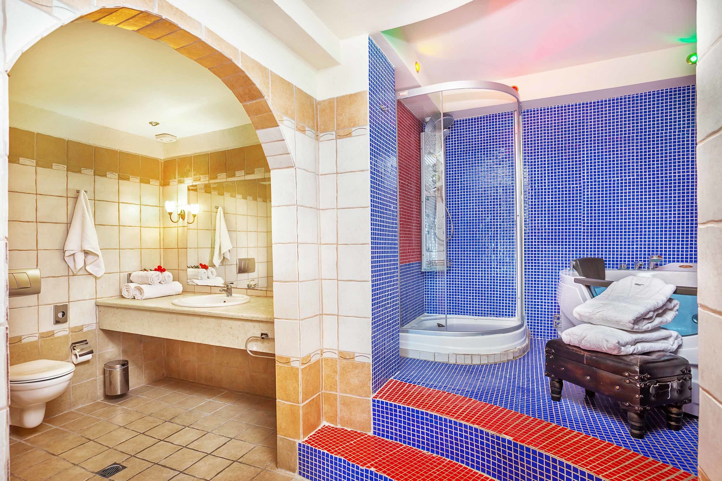 A bathroom with a beige tiled floor, a white sink, a mirror, and a blue-tiled shower area. There is a small stool with towels on it.