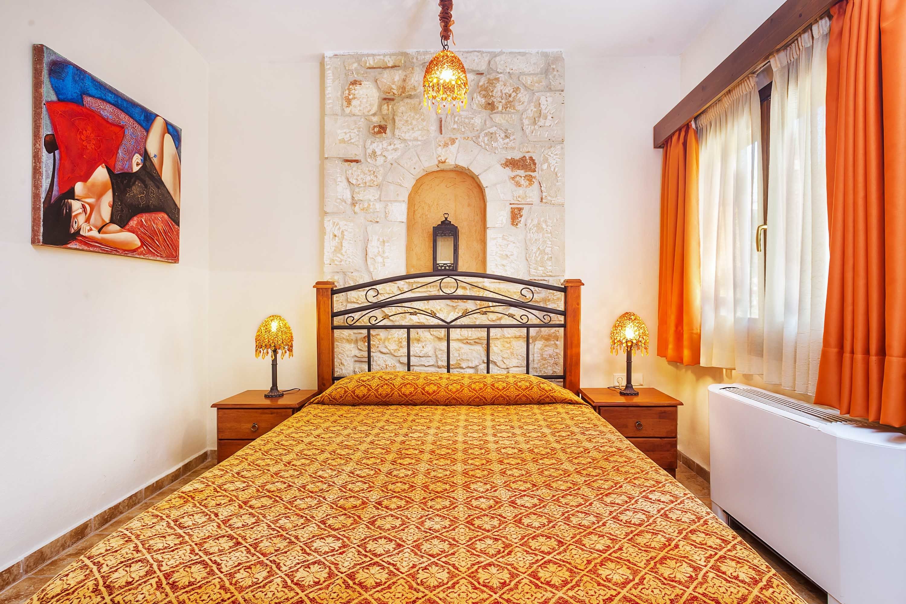 A bedroom with a patterned bedspread, a stone fireplace, two bedside lamps, and a painting on the wall.
