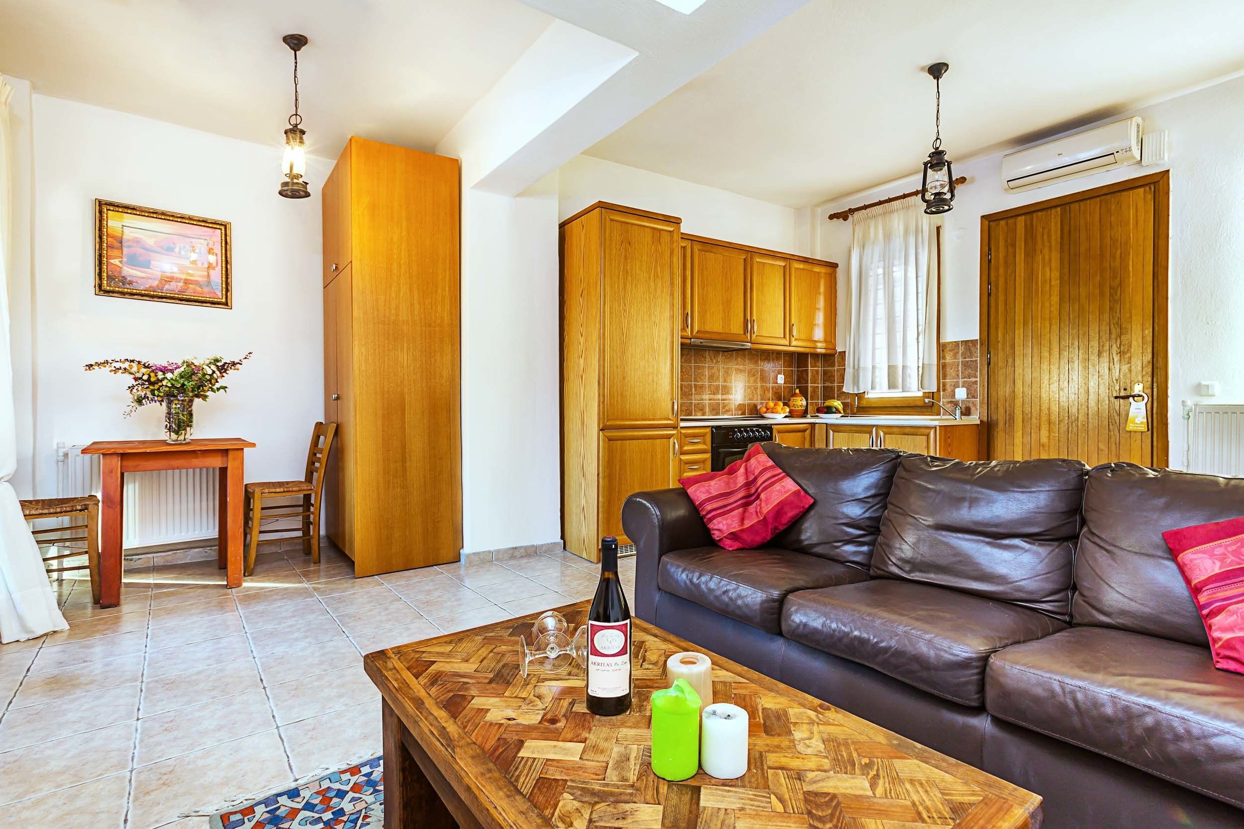 Een woonkamer met een bruine leren bank, een houten salontafel met een fles en kaarsen, en een keuken op de achtergrond met houten keukenkastjes en een hangende lamp.