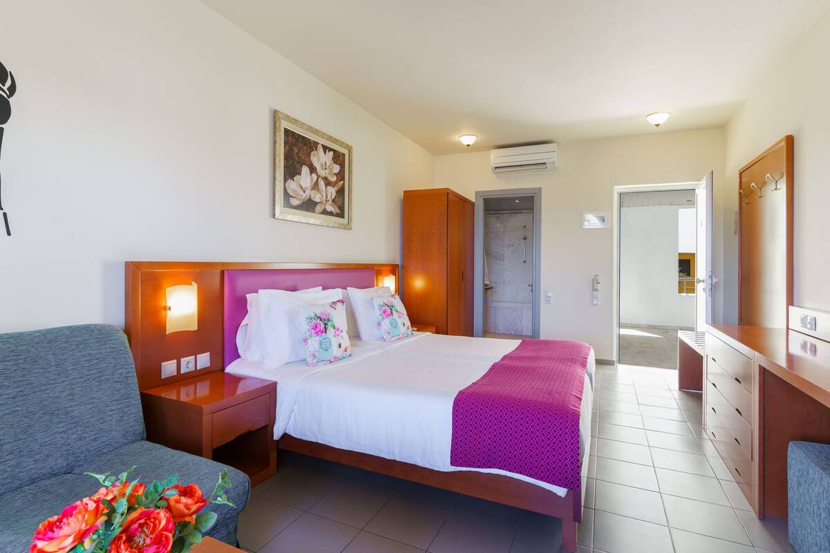 Habitación de hotel con cama, mesita de noche, cómoda, espejo y puerta que da a un baño.