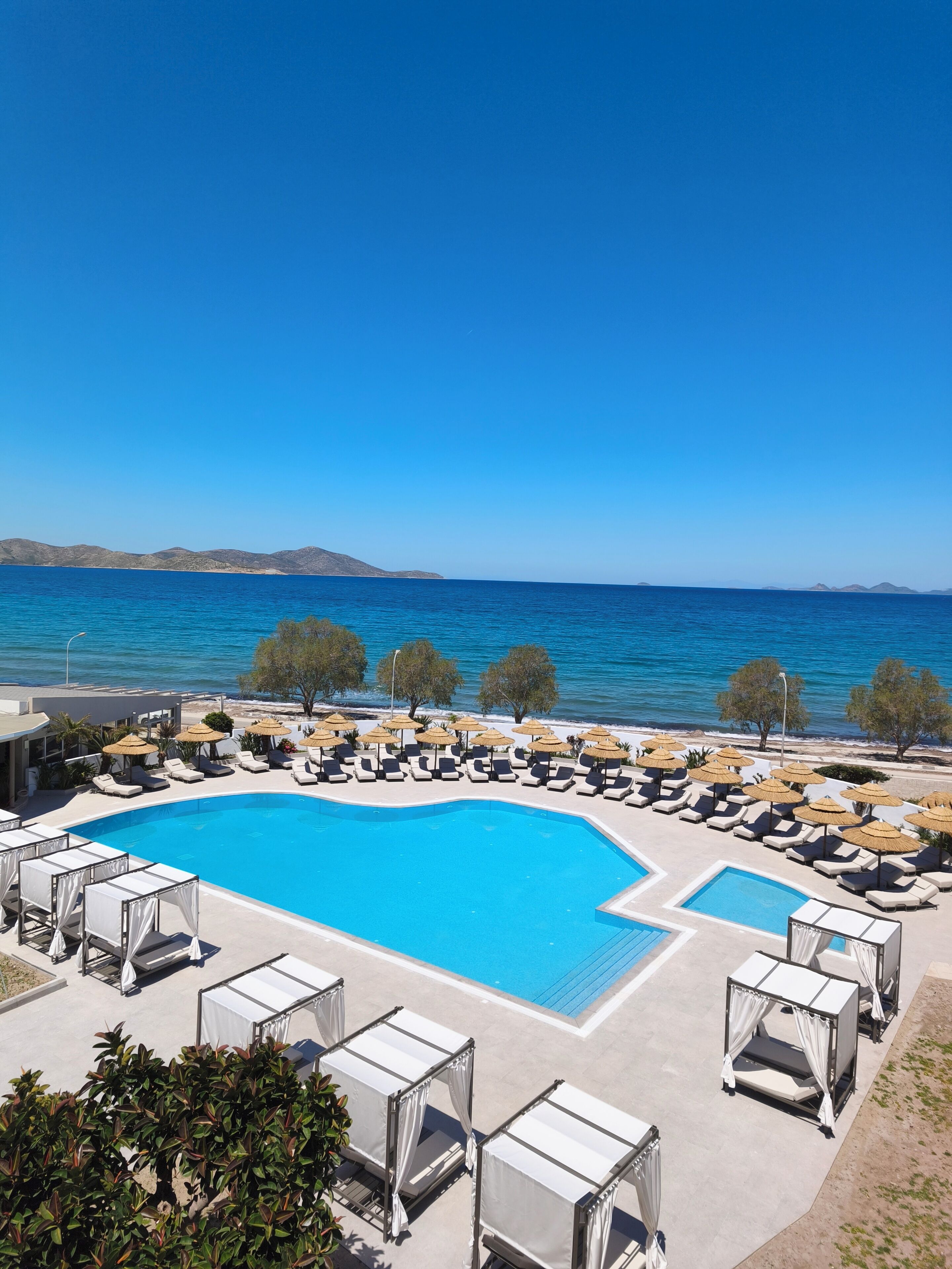 A pool area with lounge chairs and umbrellas, surrounded by a clear blue sky and a view of the sea.