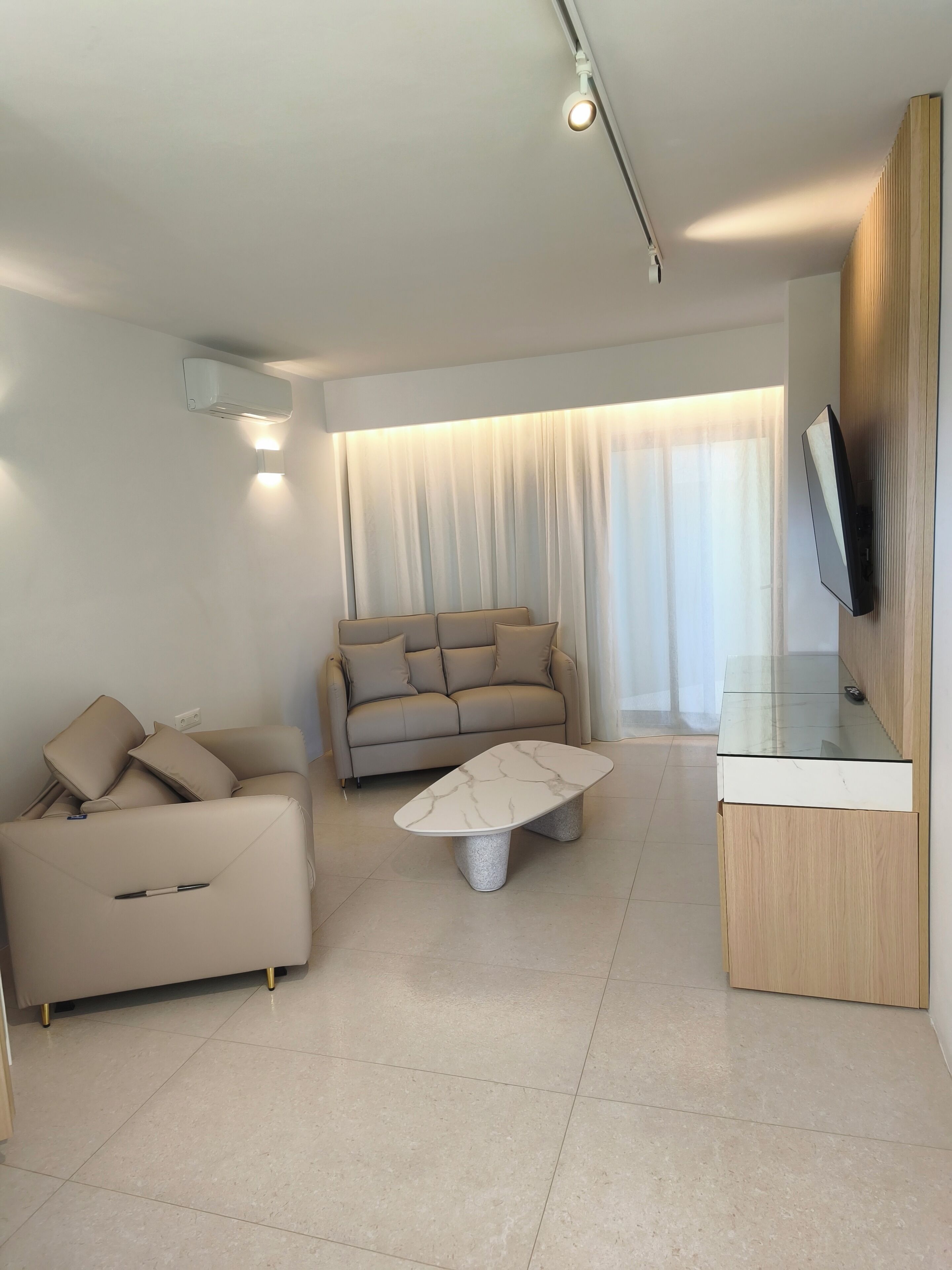 A modern living room with a sofa, a coffee table, a TV mounted on a wooden panel, and an air conditioning unit.