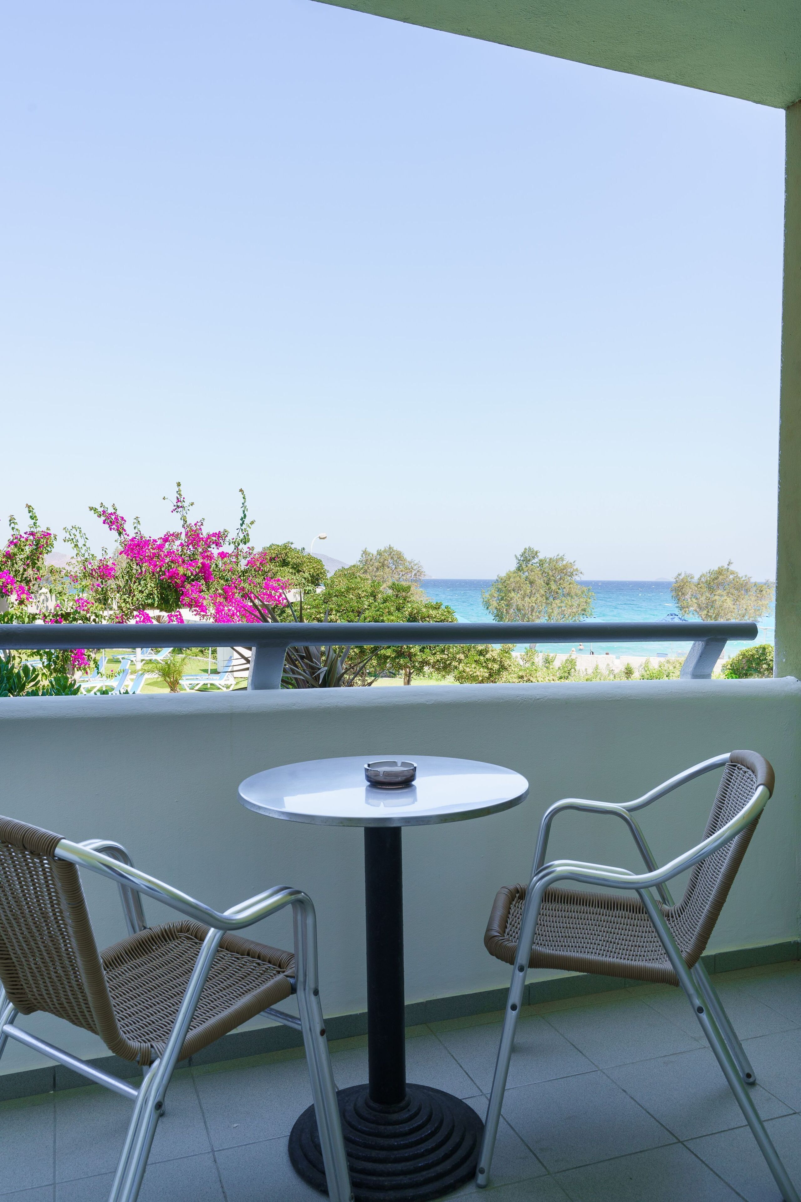 A balcony with a round table and two wicker chairs, overlooking a beach with pink flowers and the sea.