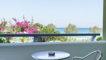 A balcony with a round table and two wicker chairs, overlooking a beach with pink flowers and the sea.