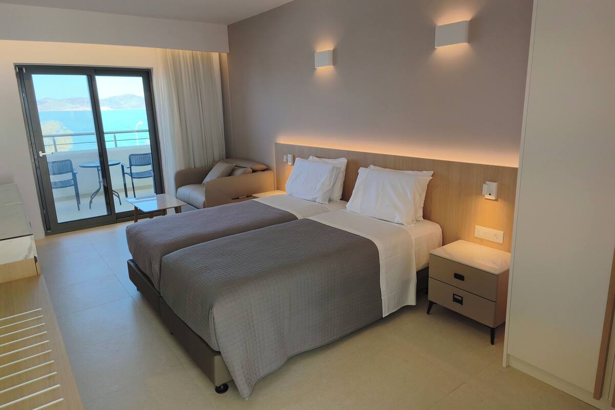 Habitación de hotel con una cama grande, una silla, una mesita y vista al mar a través de una puerta corrediza.