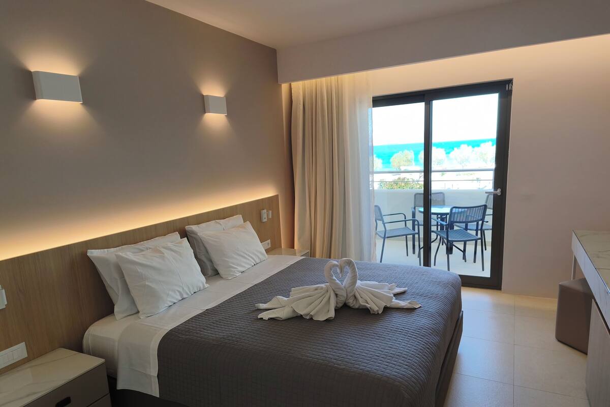 Habitación de hotel con cama, mesitas de noche, un balcón con mesa y sillas, y vista al mar.