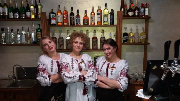 Tres personas con ropa bordada tradicional parada en una zona de bar con estantes llenos de diferentes botellas de licor al fondo.