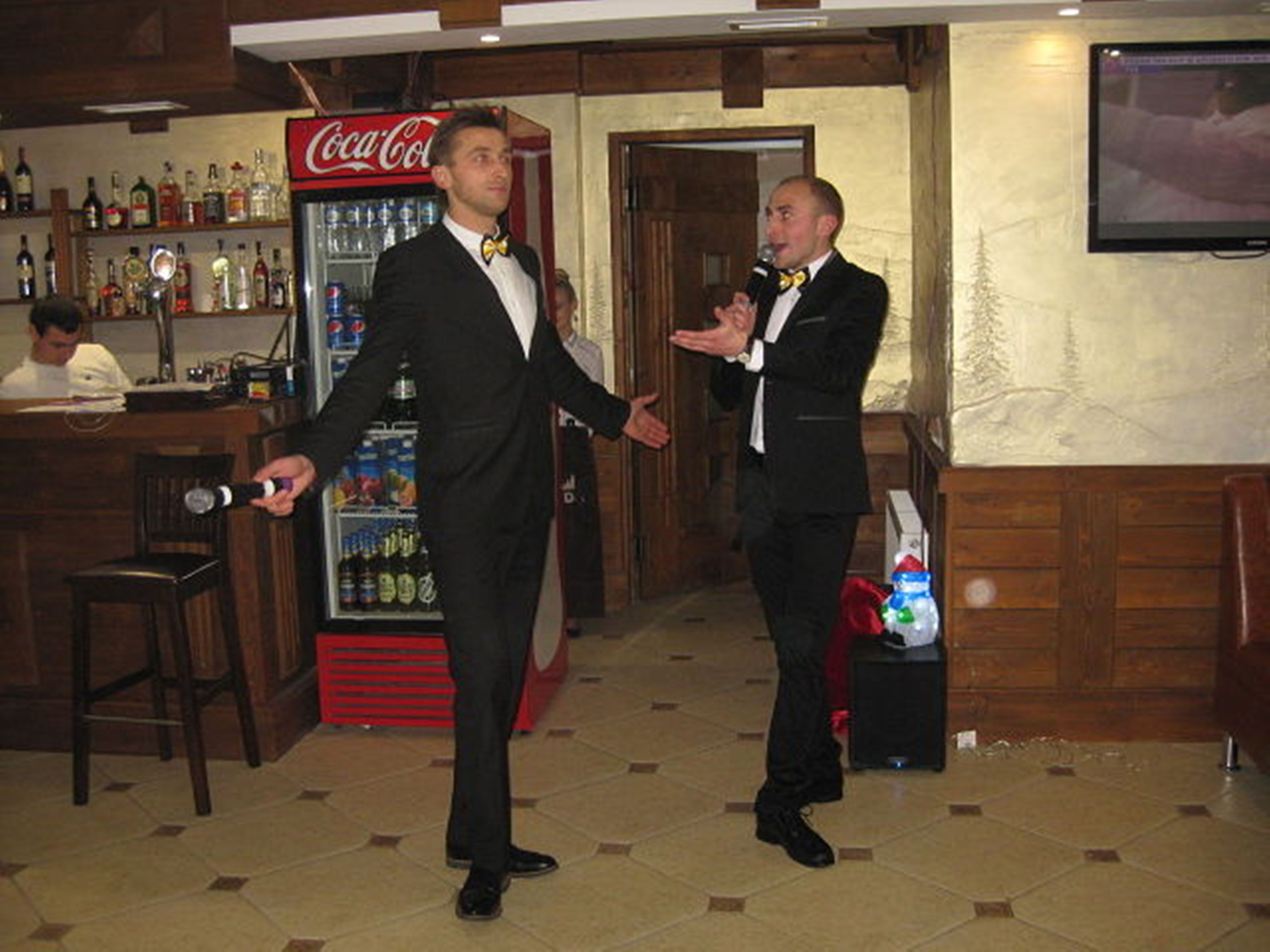 A bar area with a Coca-Cola refrigerator, a television mounted on the wall, and wooden flooring.