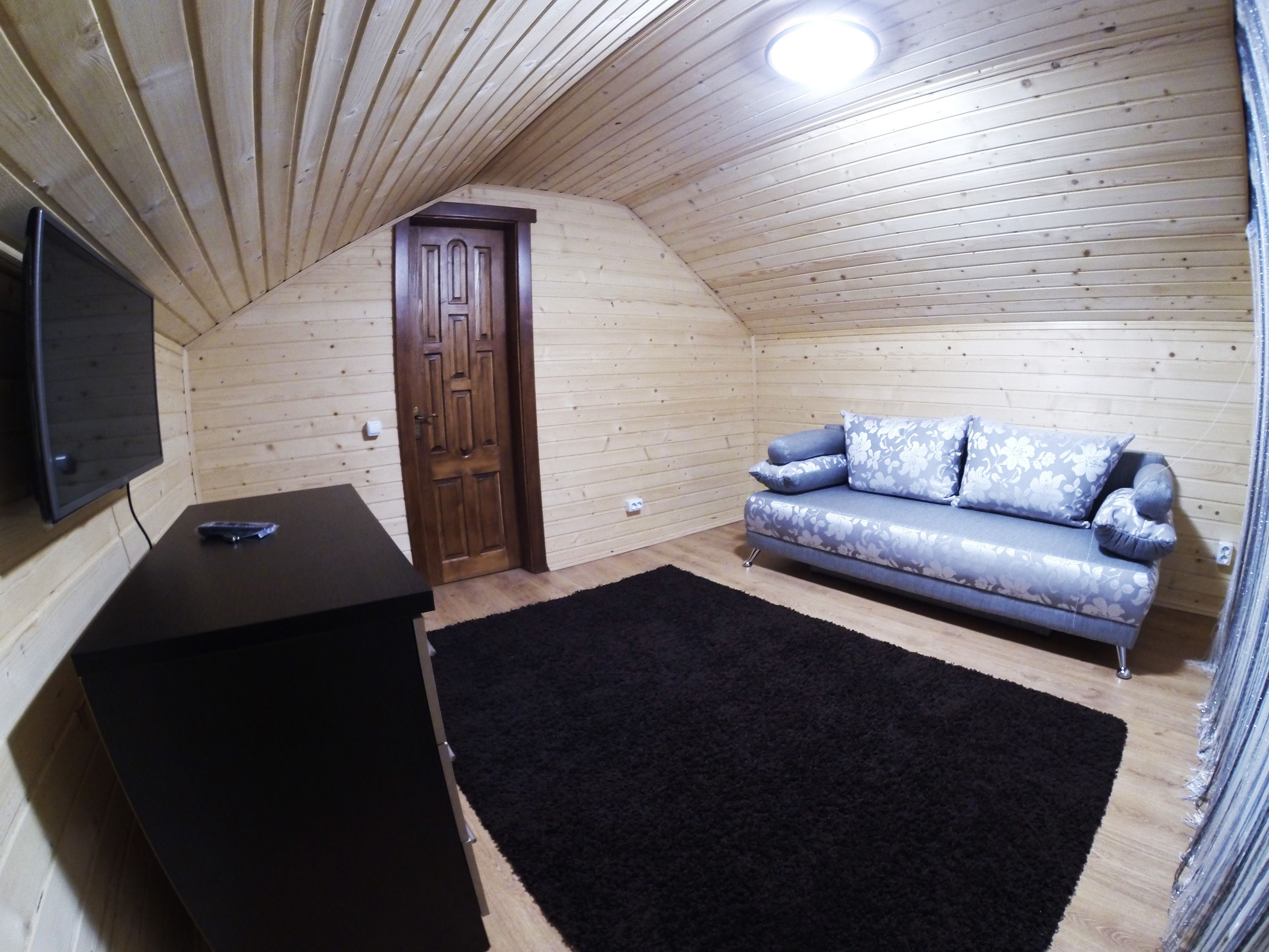 Um quarto aconchegante com teto de madeira, um sofá com almofadas florais, um tapete preto e uma TV fixada na parede.