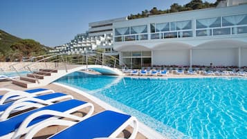 A swimming pool with blue lounge chairs, a staircase leading to a building with multiple balconies, and a clear sky.