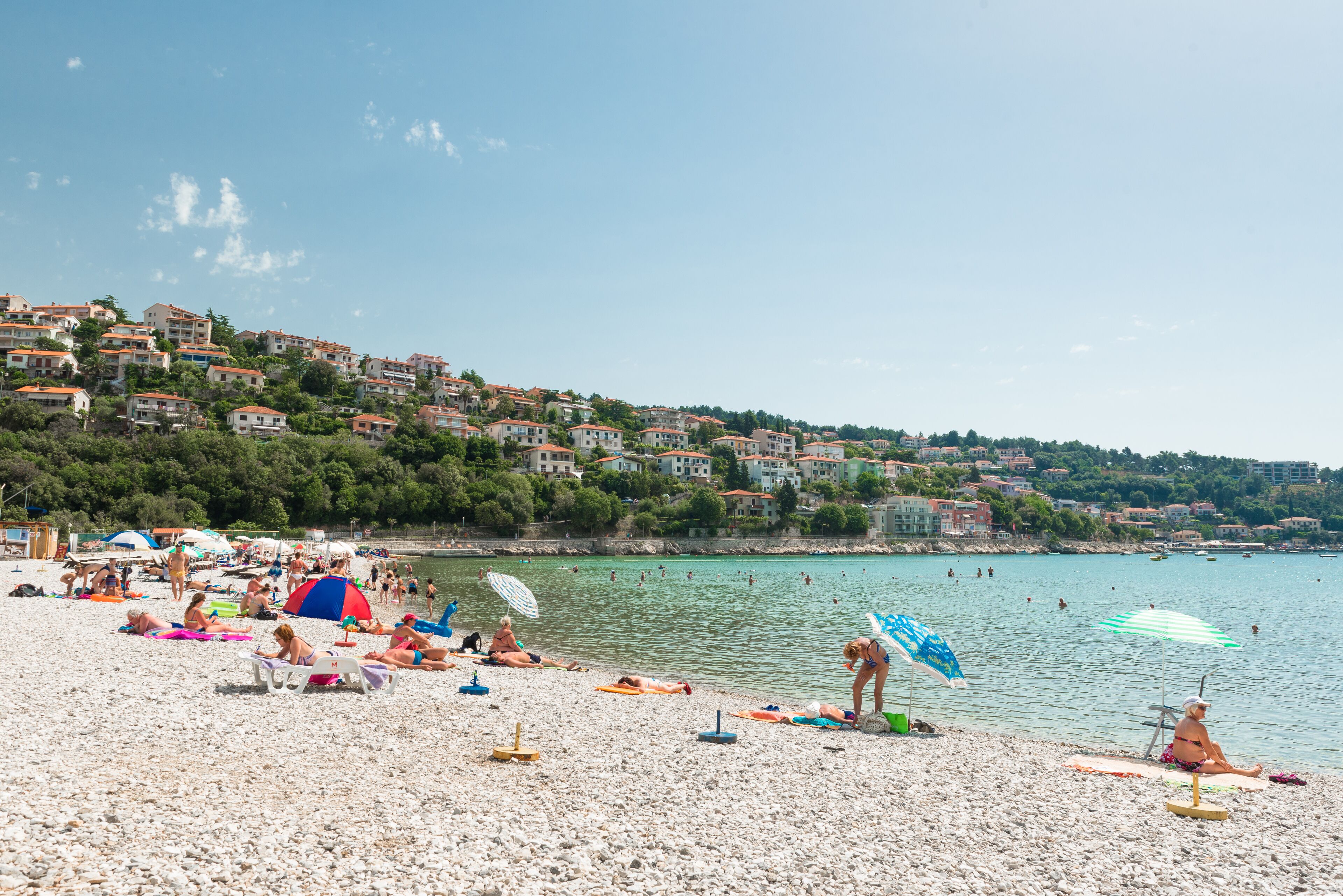 A beach with people sunbathing and swimming, surrounded by houses and a clear sky.