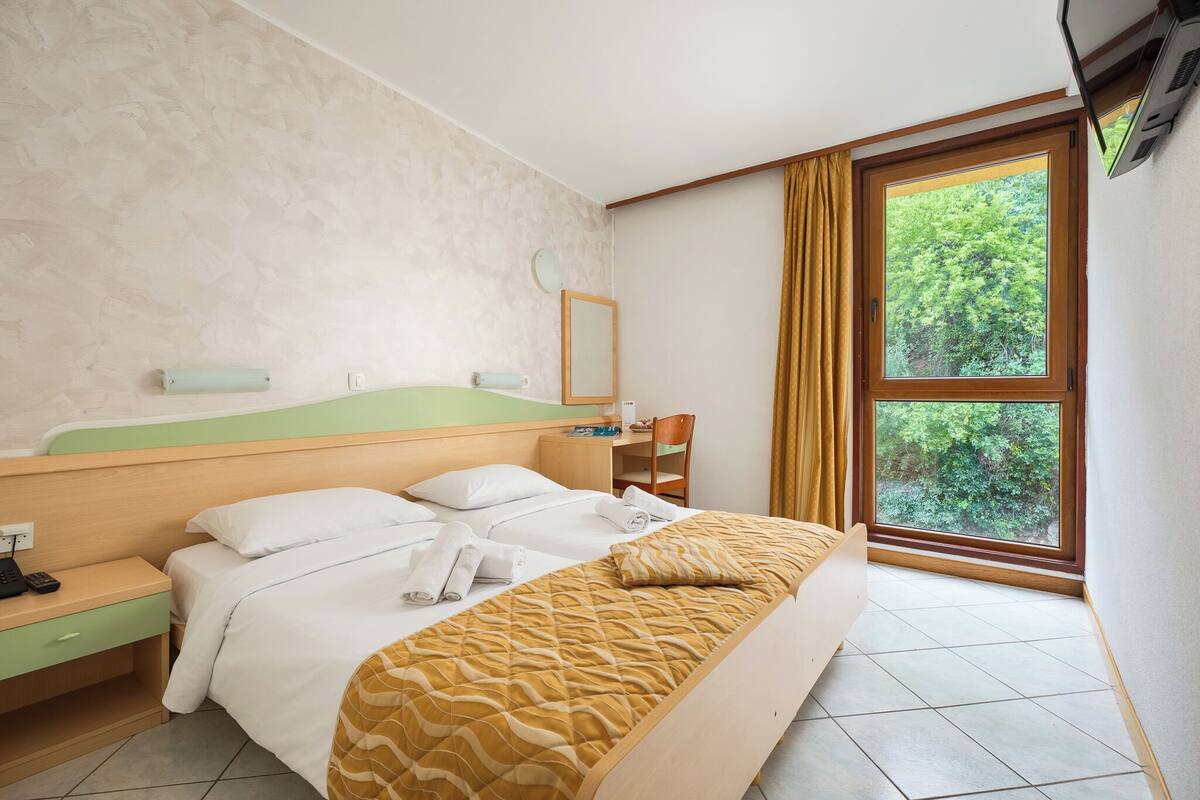 A hotel room with a bed, a desk, a chair, a window with curtains, and a wall-mounted TV.