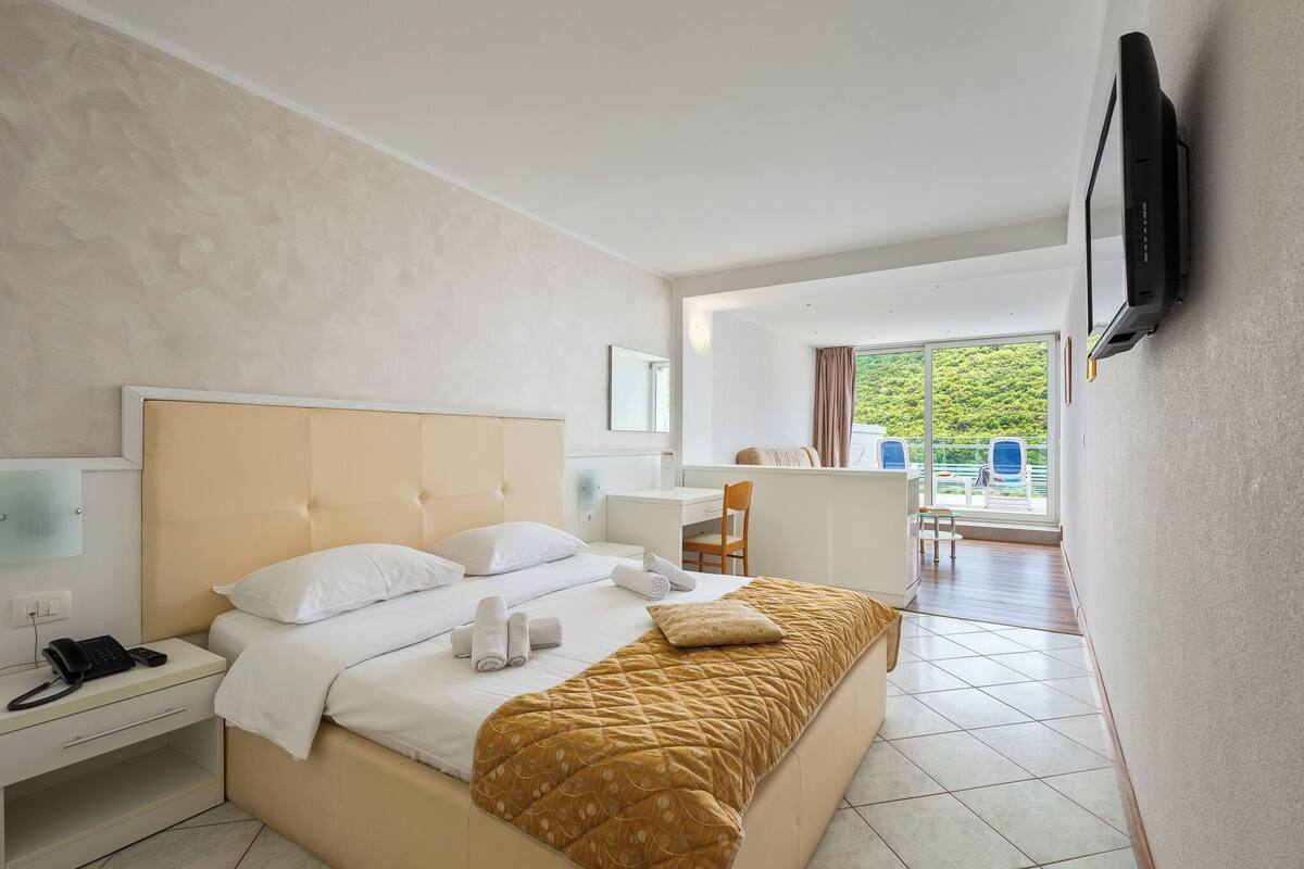 A hotel room with a bed, bedside table with a phone, a desk with a chair, a TV mounted on the wall, and a view of the outdoors through a window.