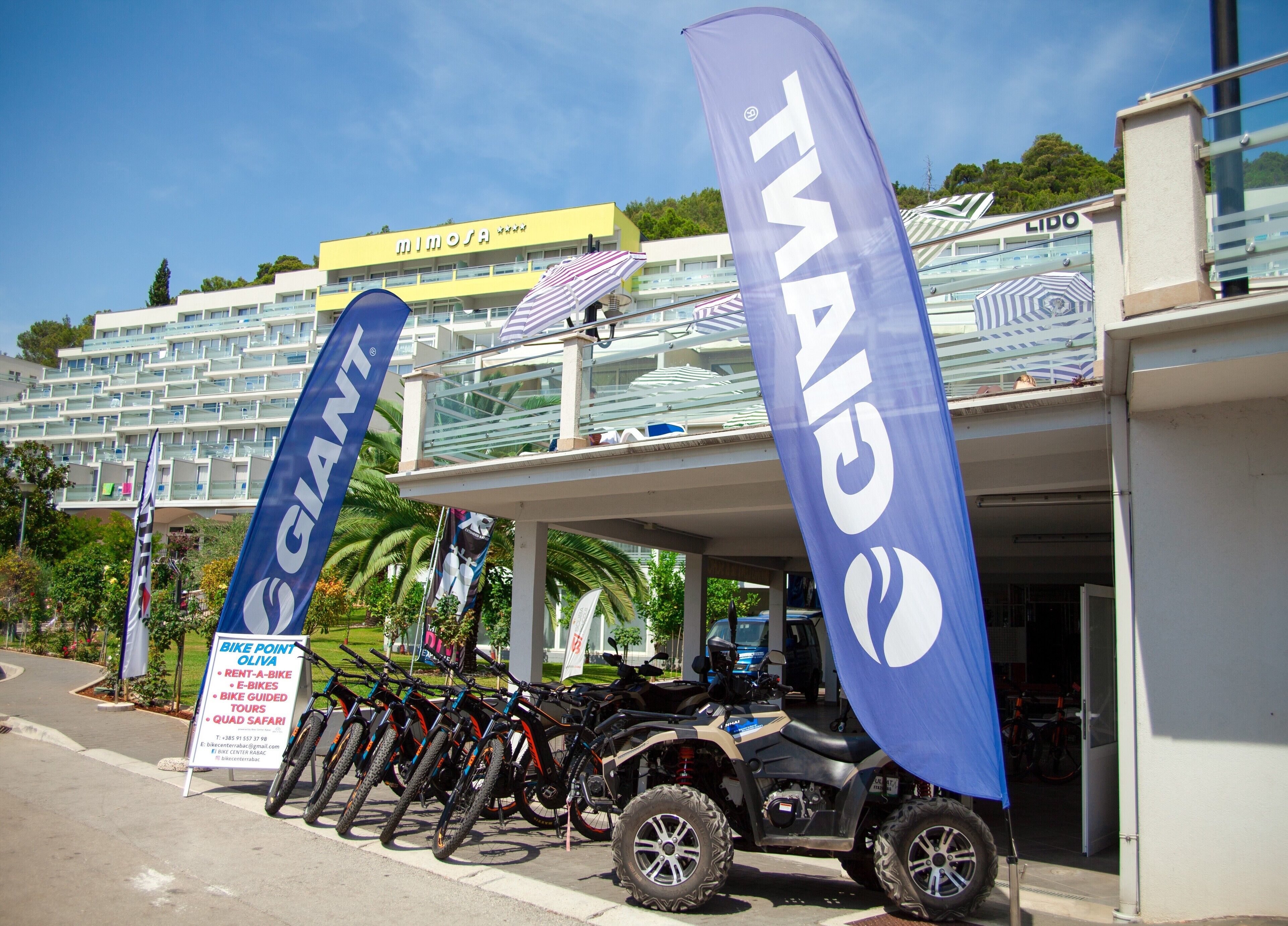 A building with multiple flags, including 'Giant' and 'TMAI', and a row of bicycles and ATVs parked in front.