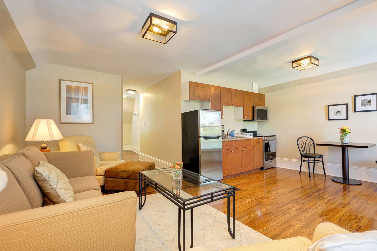 A modern living room with a glass coffee table, a sofa, and a dining area with a chair and table. A kitchen is visible in the background with wooden cabinets and a refrigerator.