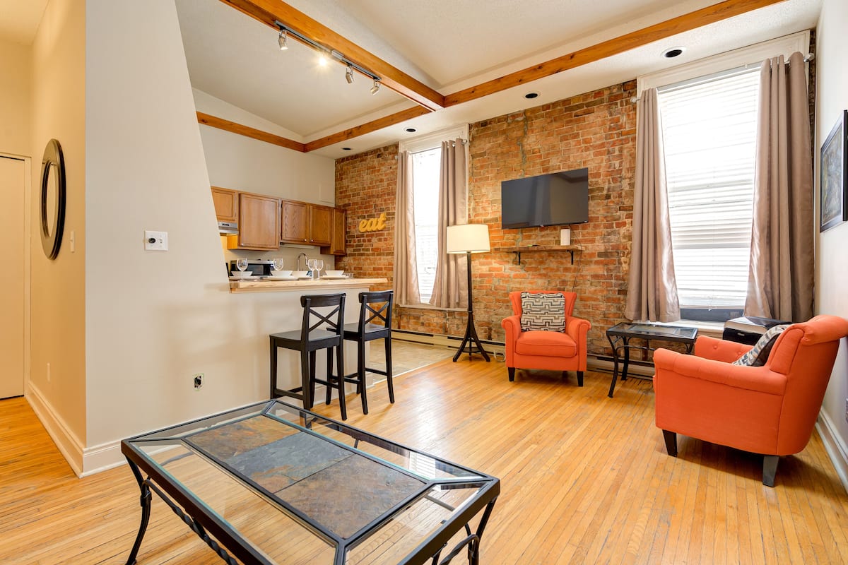 A modern living room with a glass coffee table, two armchairs, and a sofa. A kitchen with wooden cabinets and a brick wall is visible in the background.