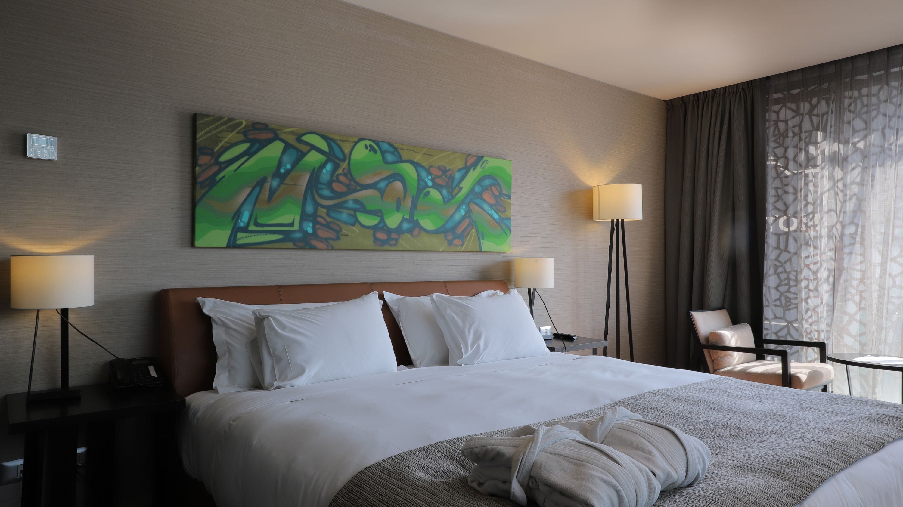 Quarto de hotel com uma cama grande, duas mesas de cabeceira, uma escrivaninha, uma cadeira, um quadro na parede e uma janela com cortinas.