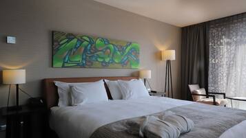 Quarto de hotel com uma cama grande, duas mesas de cabeceira, uma escrivaninha, uma cadeira, um quadro na parede e uma janela com cortinas.