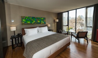 Habitación de hotel con una cama grande, dos mesitas de noche con lámparas, una silla, una pequeña mesa y un ventanal con vistas a la ciudad.