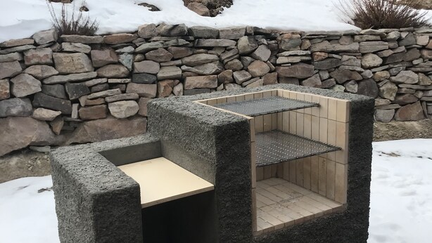 Un banco de piedra con una reja metálica y un asiento de madera, al aire libre, con un muro de piedra y nieve al fondo.