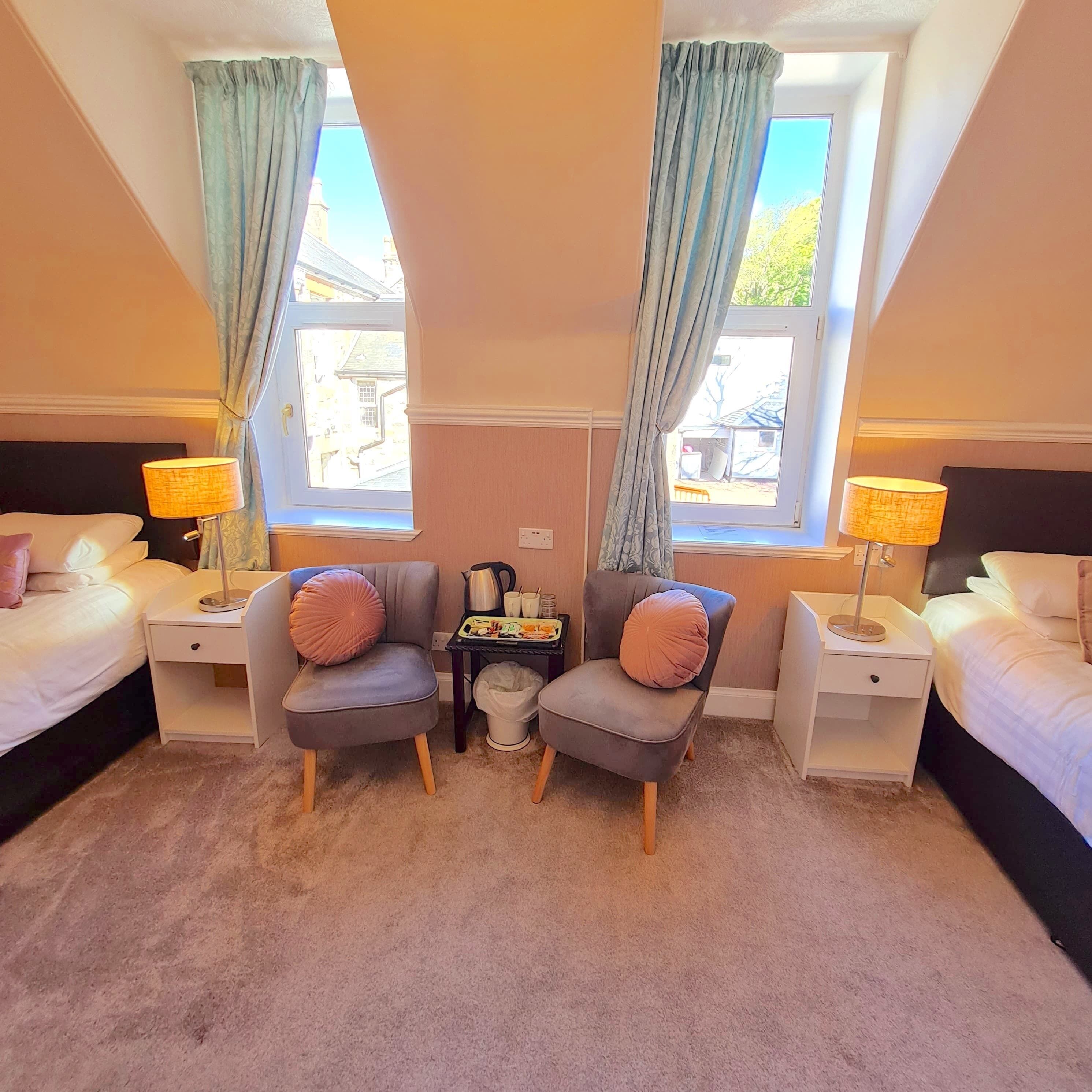 A hotel room with two single beds, a small table with a kettle, two armchairs with cushions, and two bedside lamps.
