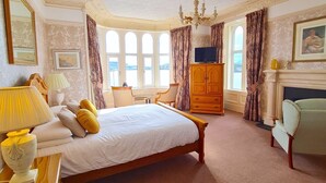 A spacious bedroom with a large bed, a wooden wardrobe, a chandelier, and a fireplace. - Corriemar Guest House (Oban)