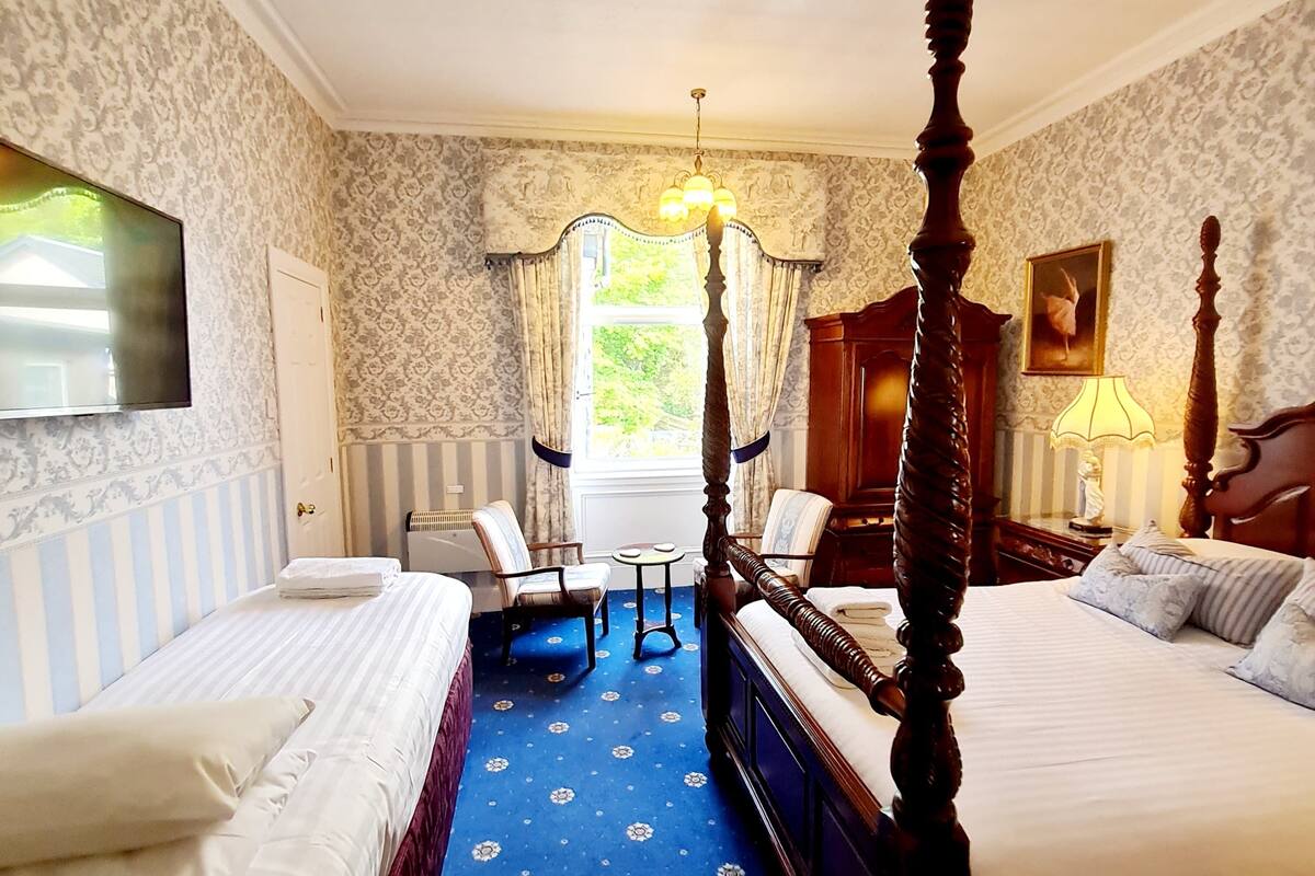 A hotel room with two beds, a large window with curtains, a chandelier, and a painting on the wall.