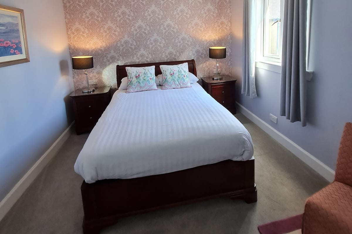 A bedroom with a large bed, two bedside tables with lamps, a patterned wallpaper, a window with curtains, and a carpeted floor.