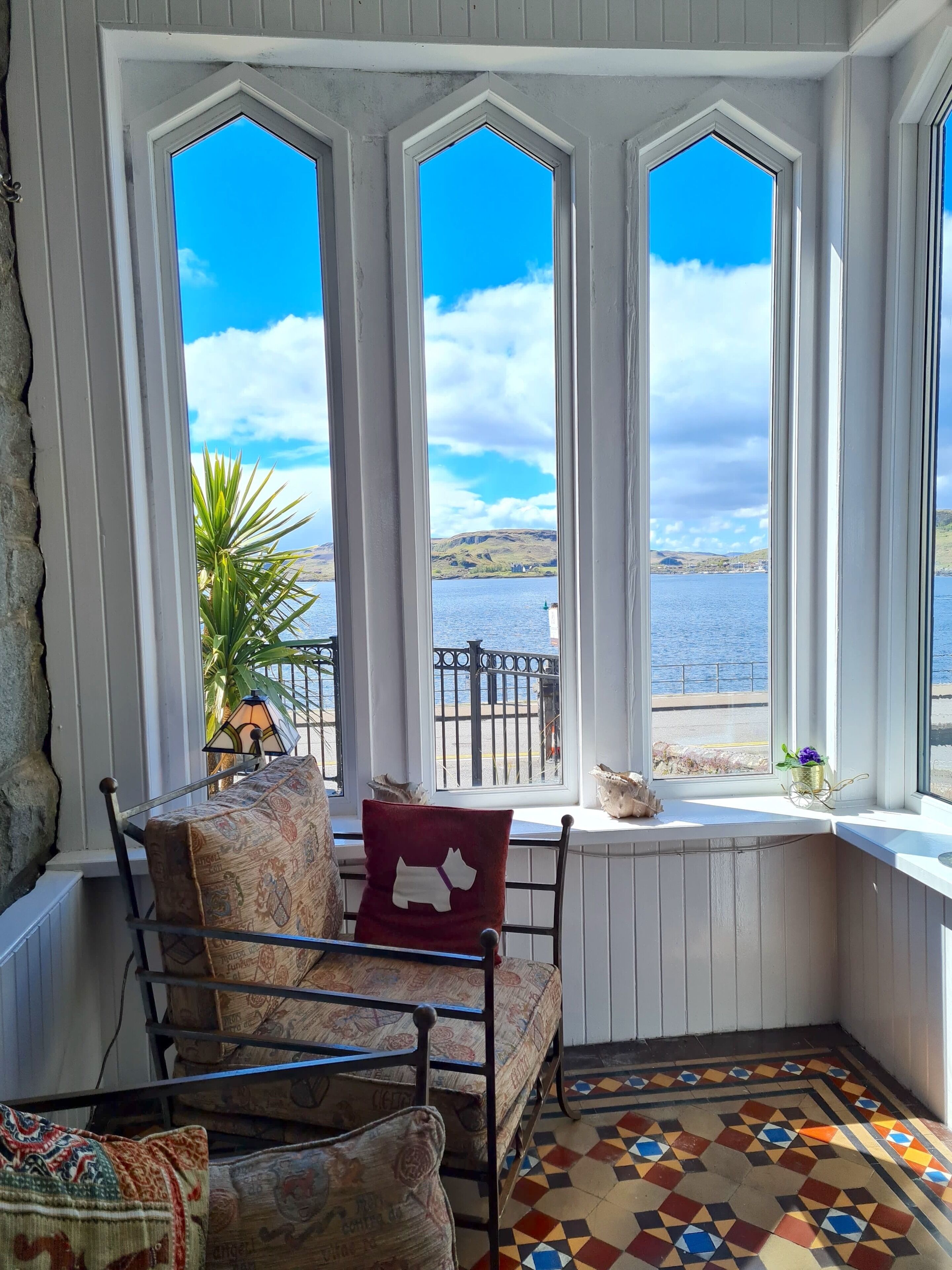 A cozy room with a view of a lake and hills. It features a metal-framed chair, a patterned rug, and a window with a view of the water.