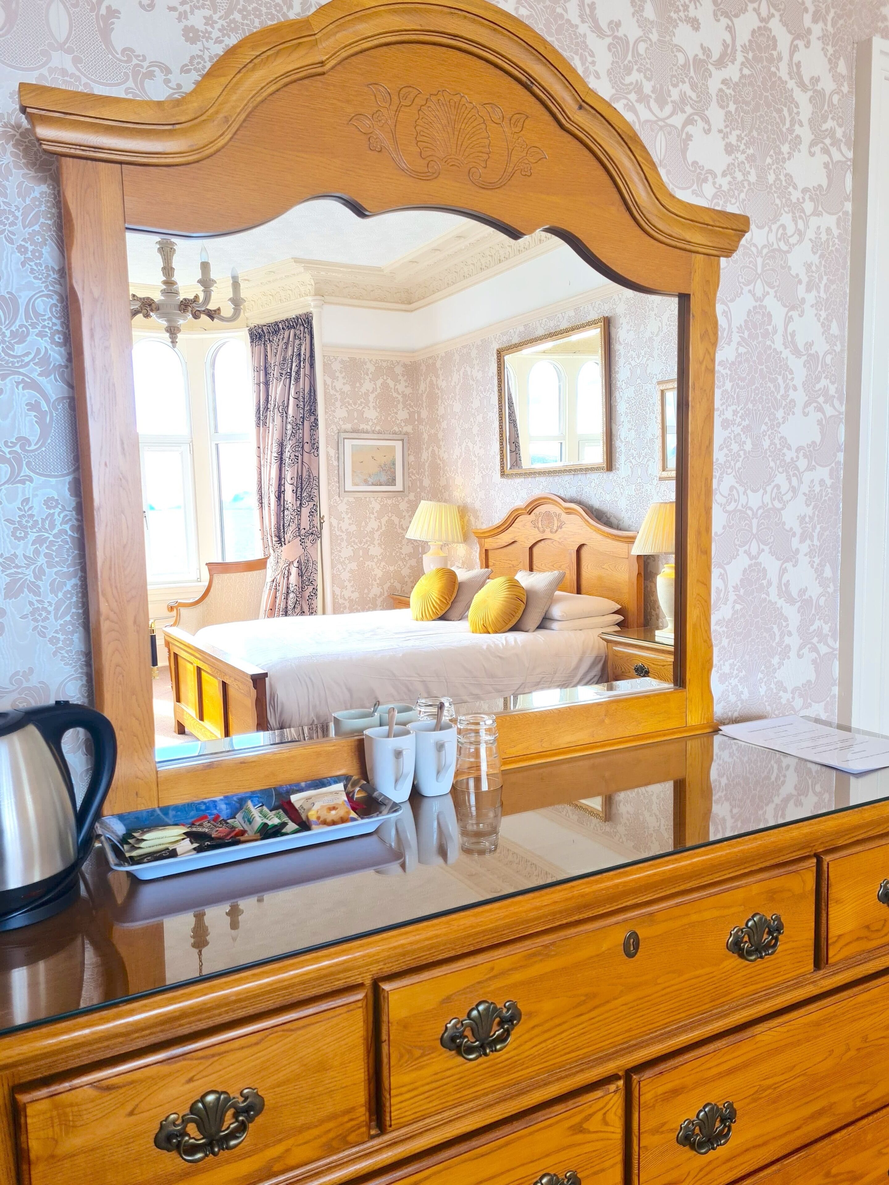 A bedroom with a large mirror, wooden furniture, a bed with yellow pillows, and a bedside table with a kettle and a tray of snacks.
