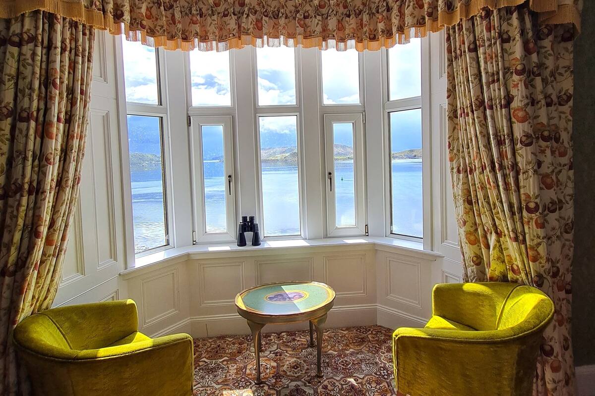 A room with a bay window, two yellow armchairs, a small table, patterned carpet, and curtains with a fruit pattern.