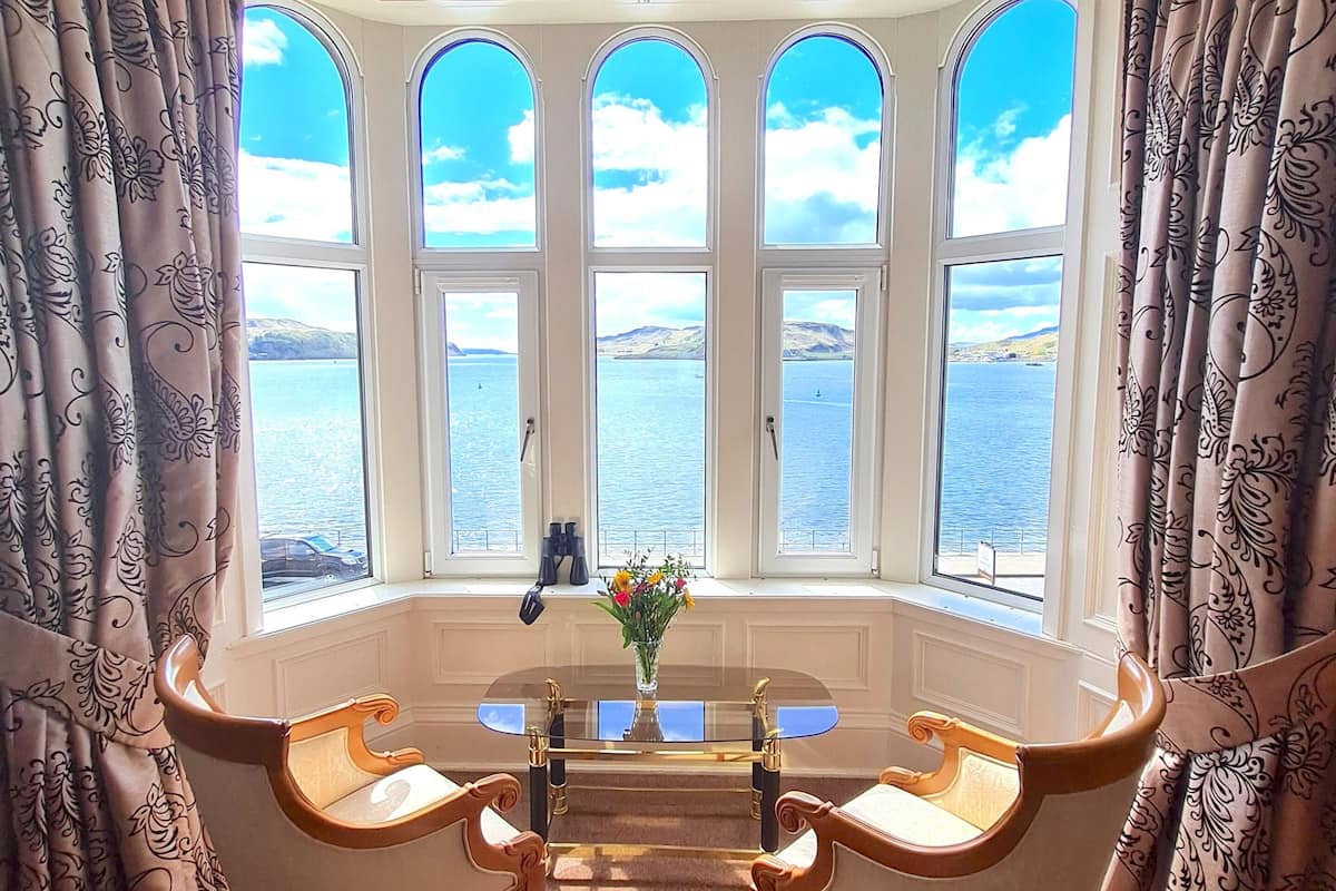 A room with a bay window overlooking a body of water, a table with chairs, and a vase with flowers.