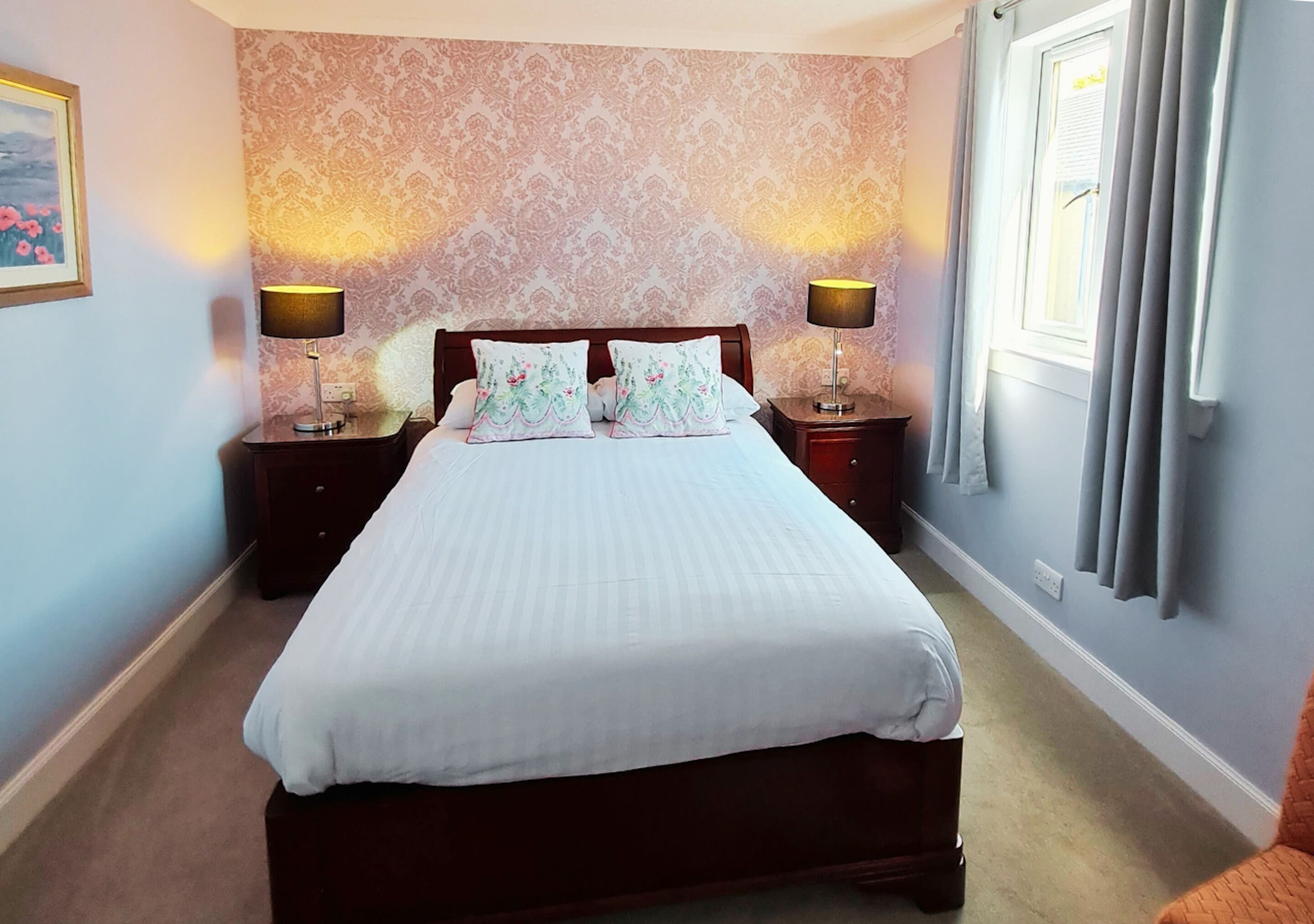 A neatly made bed with a white comforter and two decorative pillows, flanked by two bedside tables with lamps, in a room with patterned wallpaper and a window with curtains.