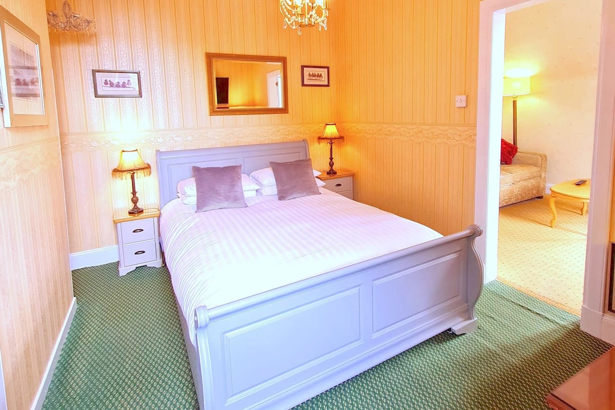 A bedroom with a large bed, two bedside tables with lamps, a chandelier, a mirror, and a door leading to another room.