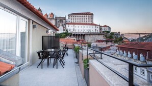 A balcony with outdoor seating, overlooking a historic building with red-tiled roofs and a large white structure with arched windows. - The Editory House Ribeira Porto Hotel (Porto)