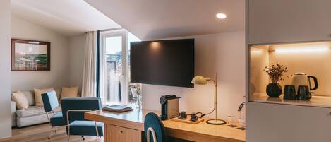 A modern room with a desk, chairs, a flat-screen TV, a coffee maker, and a view of the outdoors.