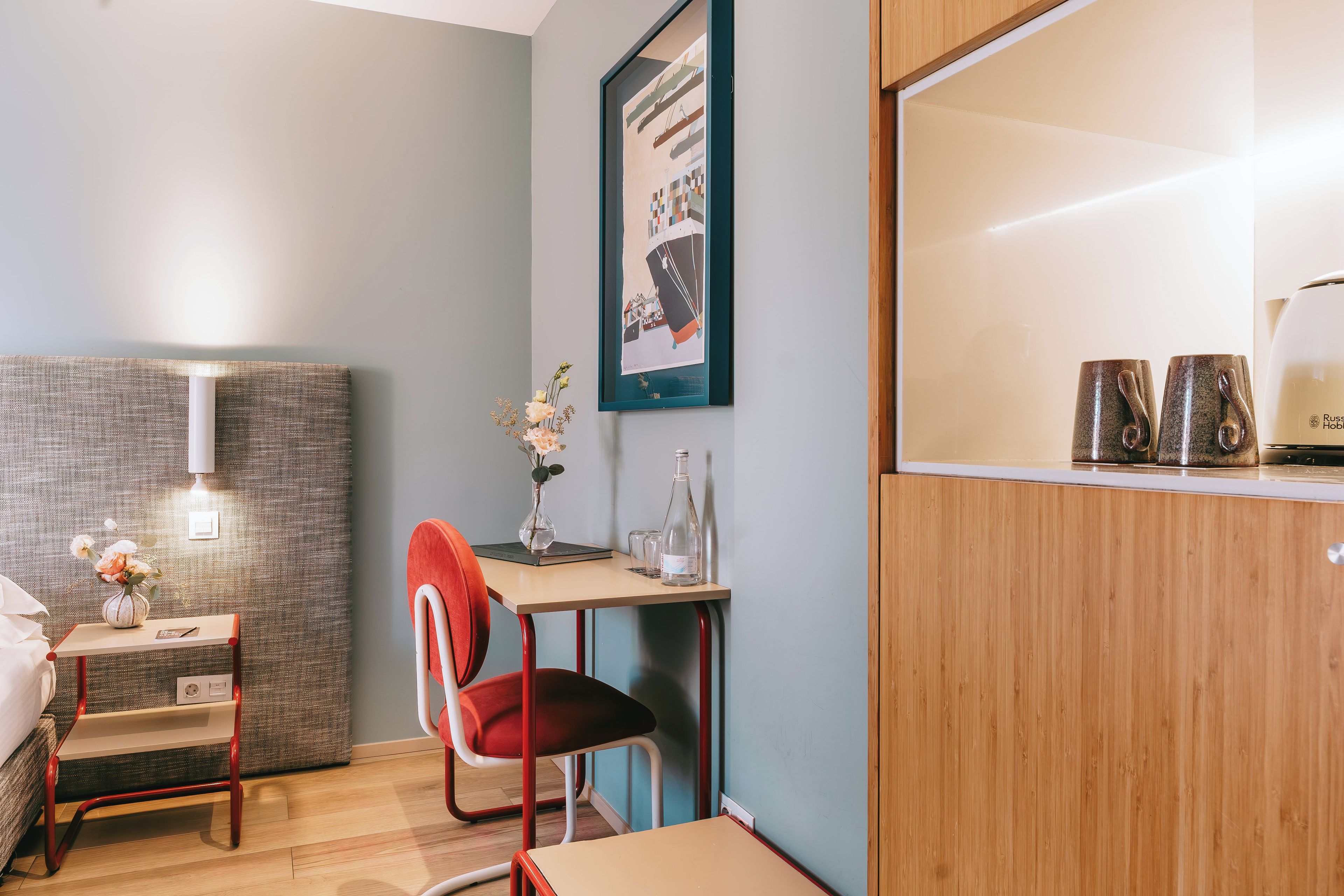 A modern hotel room with a bed, a desk, a chair, a vase with flowers, a framed picture, and a coffee machine.
