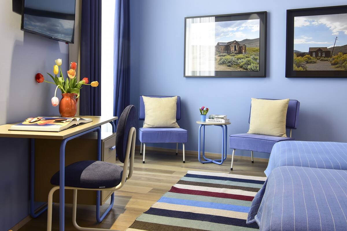 A hotel room with a desk, two chairs, a small table with a vase of flowers, two beds, a striped rug, and two framed pictures on the wall.
